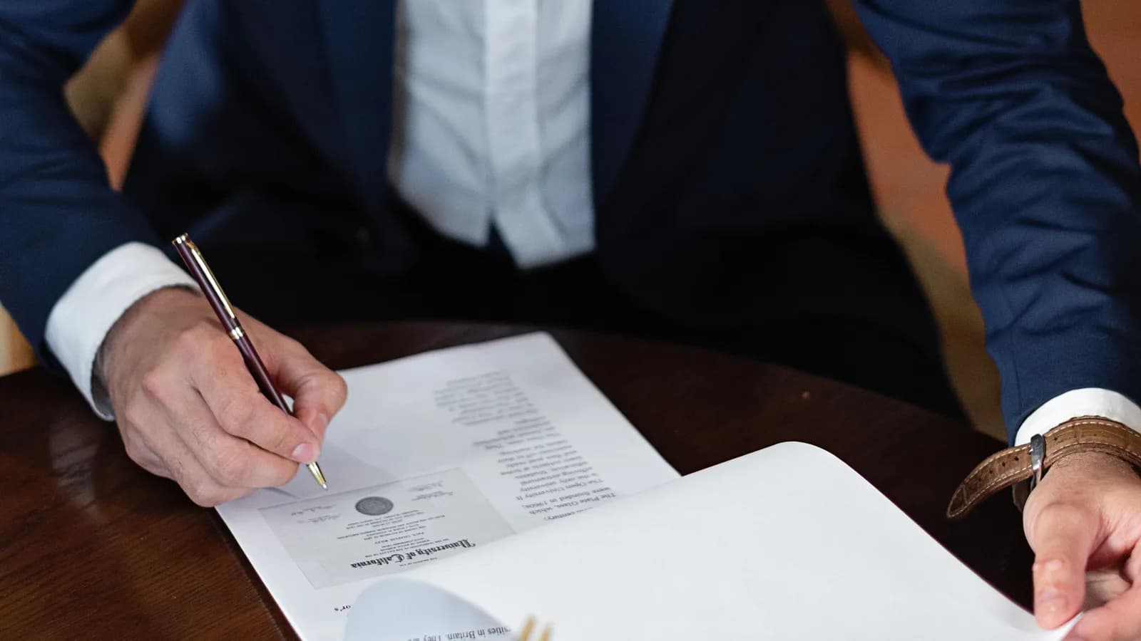 A person in a suit signing an official certificate-style document at a desk