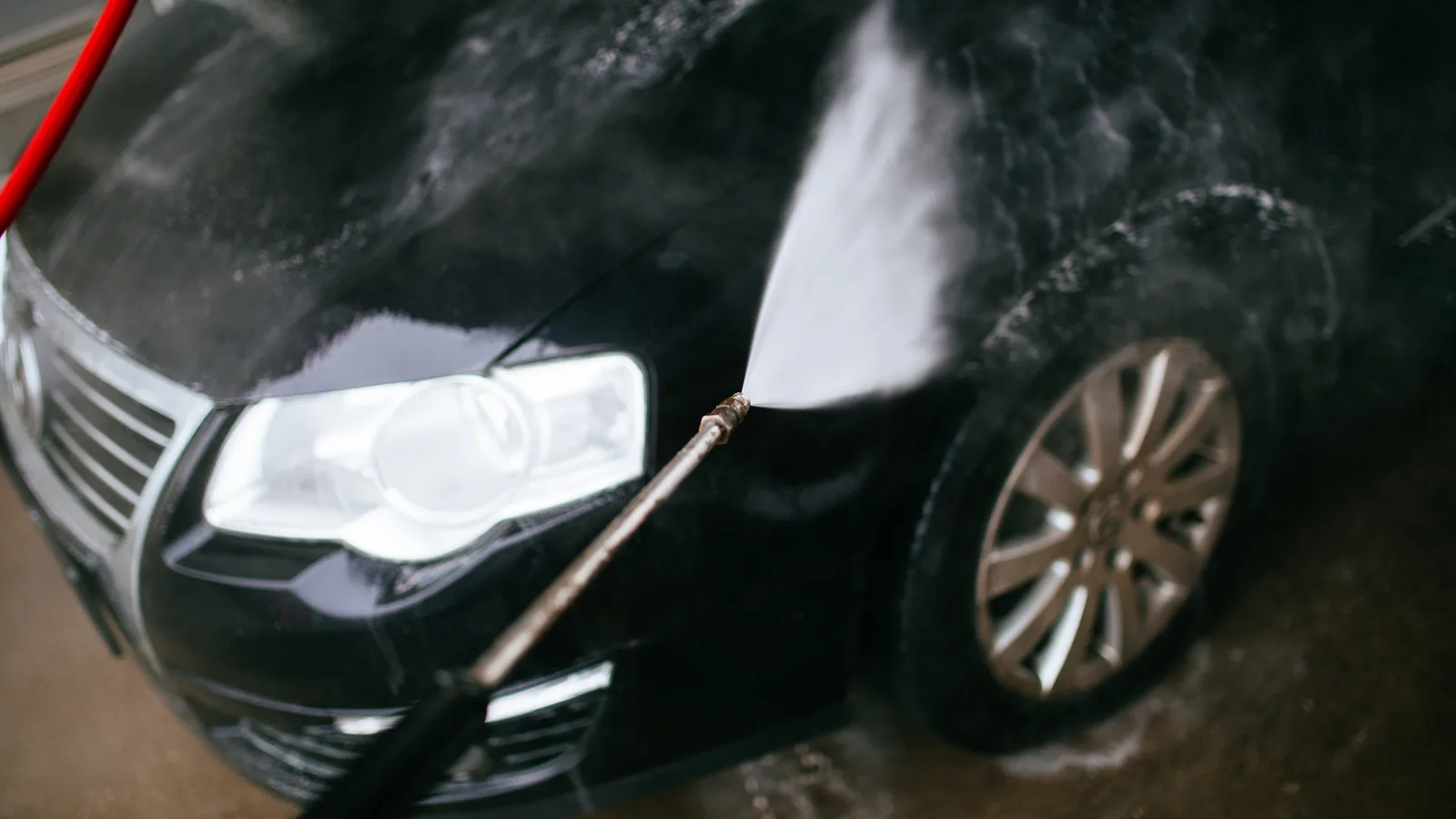 A black car being pressure-washed, with foam covering the front bumper, headlight and wheel