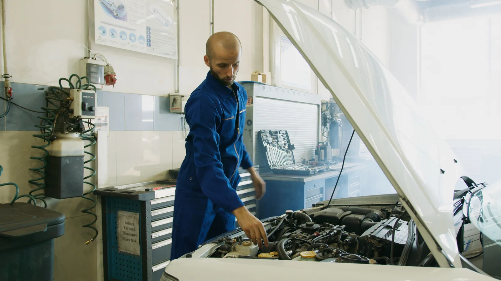 A mechanic in blue overalls standing beside a white car with its bonnet raised in a workshop