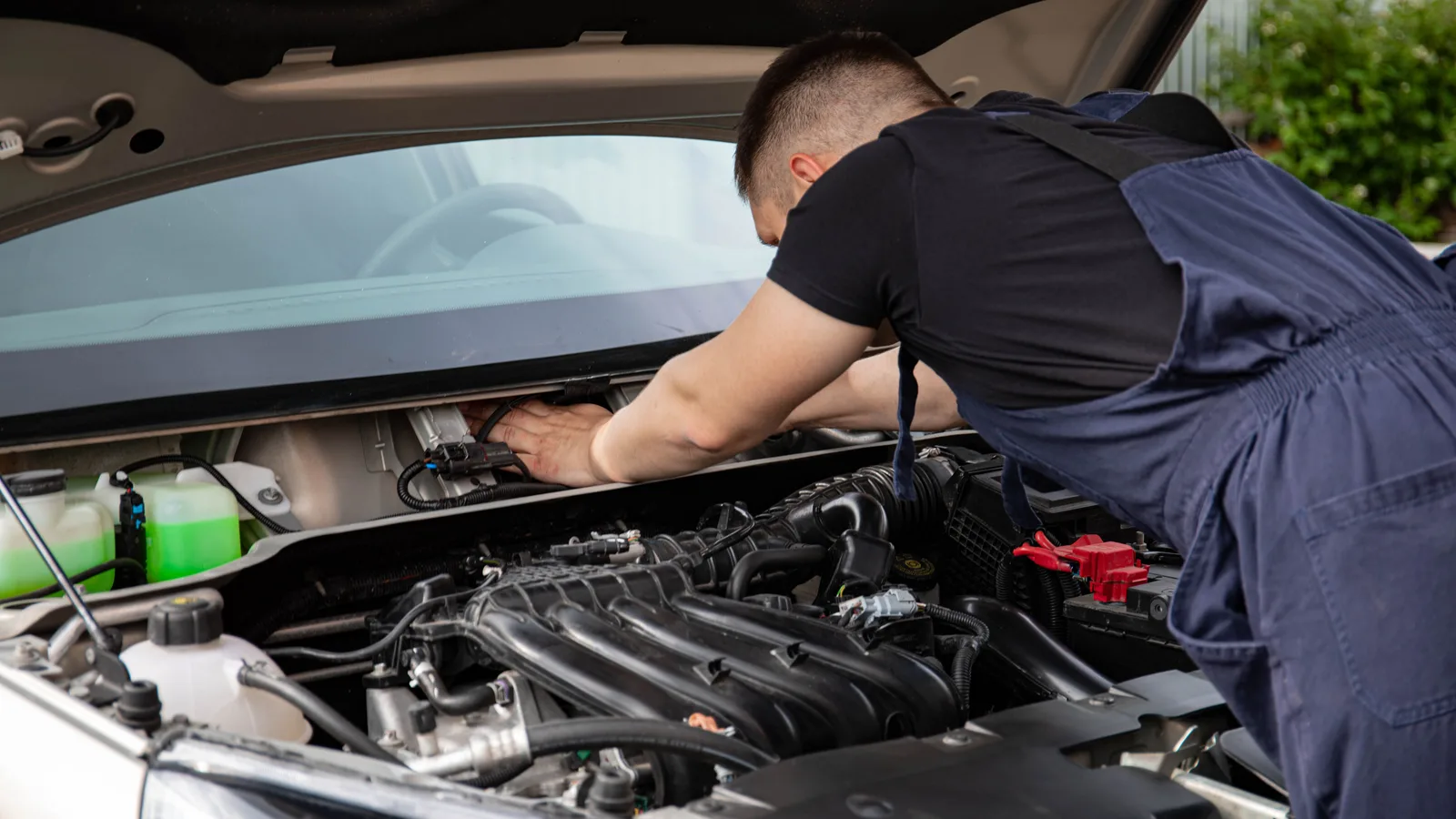 A mechanic in overalls leaning into the open bonnet of a silver car to work on the engine