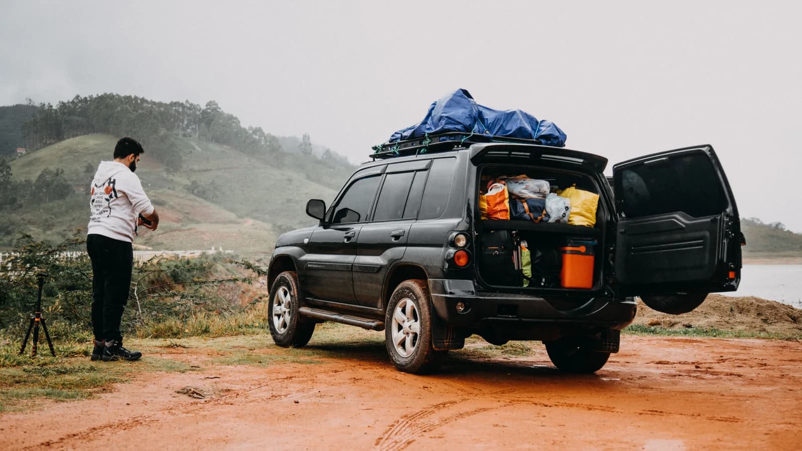 An SUV with its boot open and packed with bags and camping equipment on a dirt track beside a lake