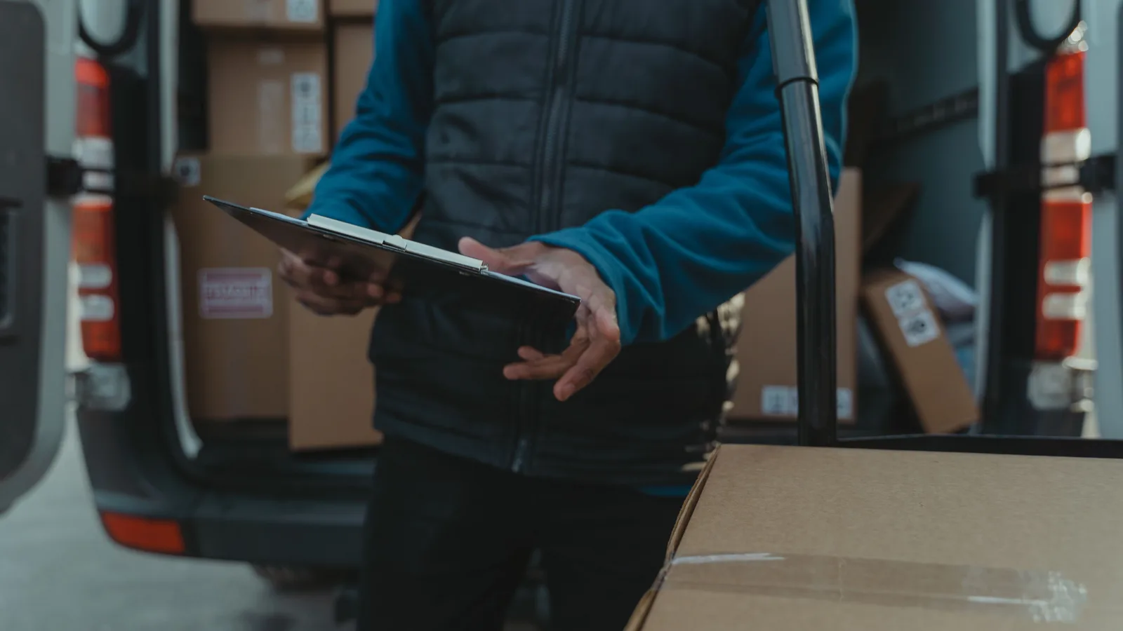 A driver holding a clipboard beside the open rear doors of a van loaded with parcels