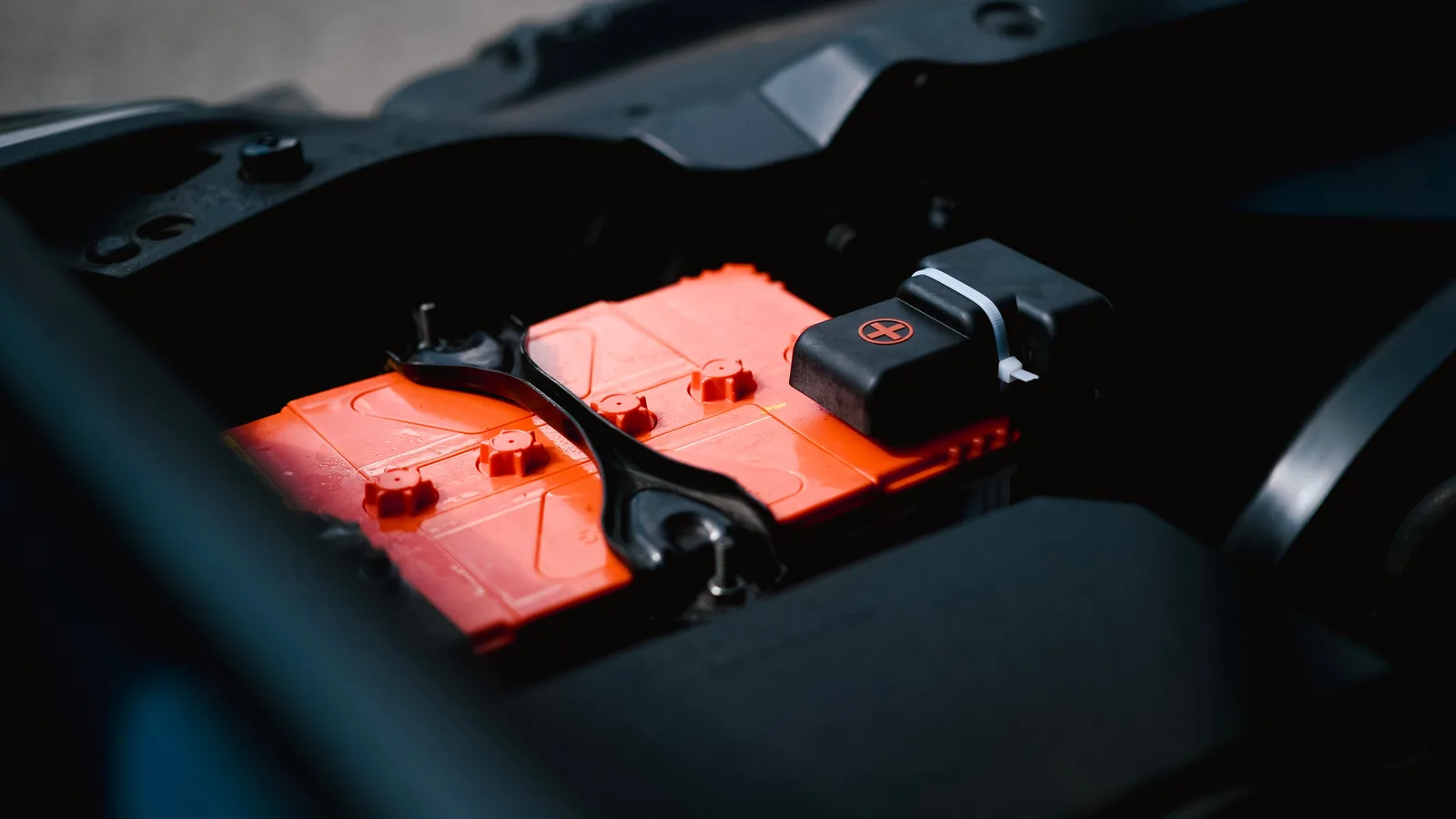 A close-up of a red lead-acid battery mounted in a vehicle, positive terminal visible