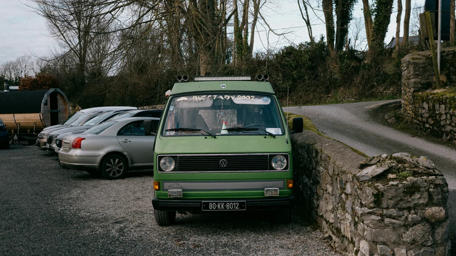 A green camper van parked on gravel beside a stone wall and a quiet country road