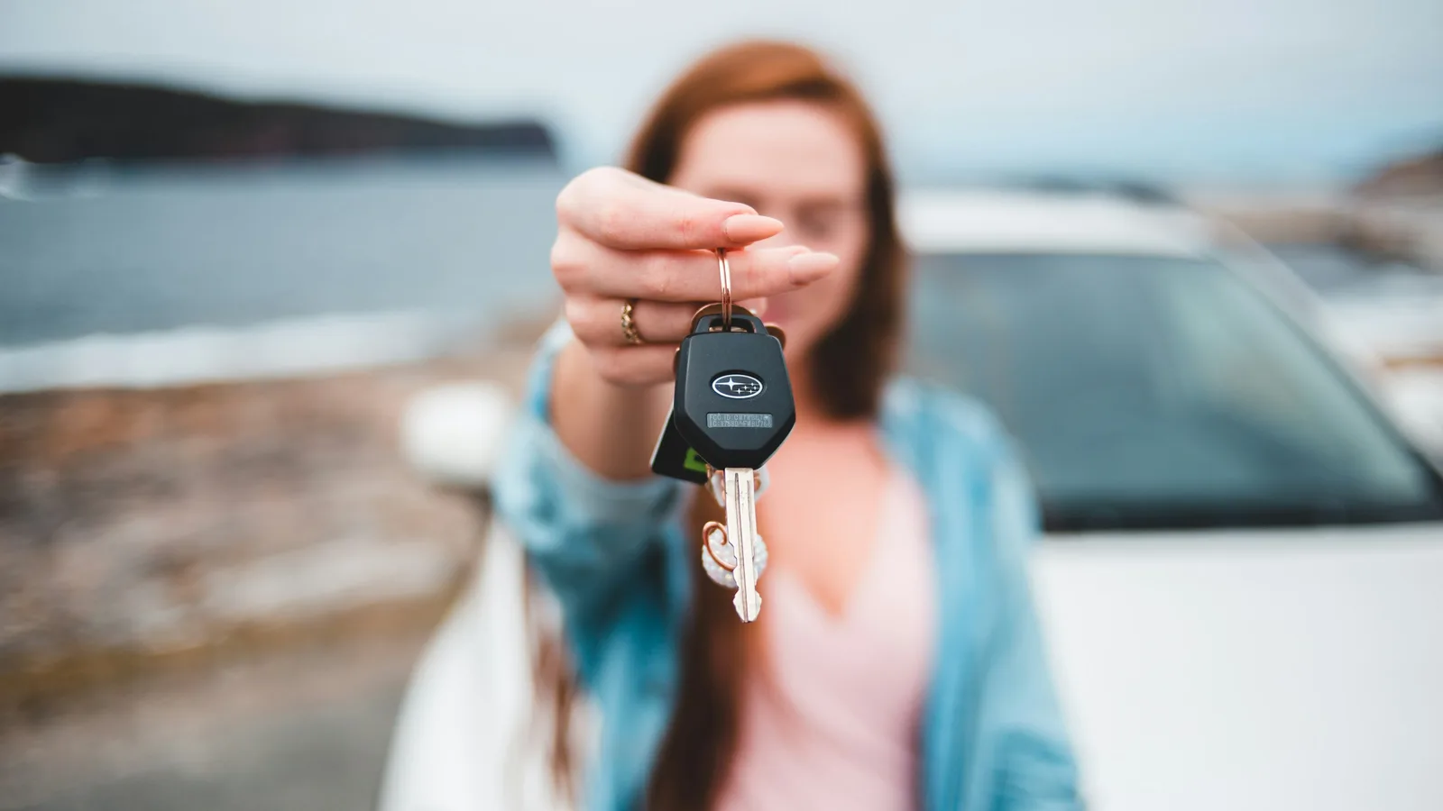 A woman holding out a Subaru key fob toward the camera in front of a white car on a coastal roadside, face blurred