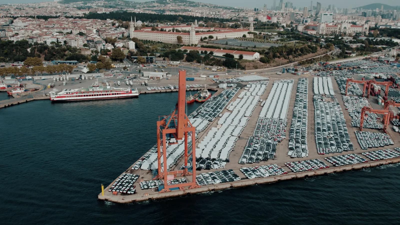 Port terminal from above, imported vehicles in rows with cranes and a ferry behind