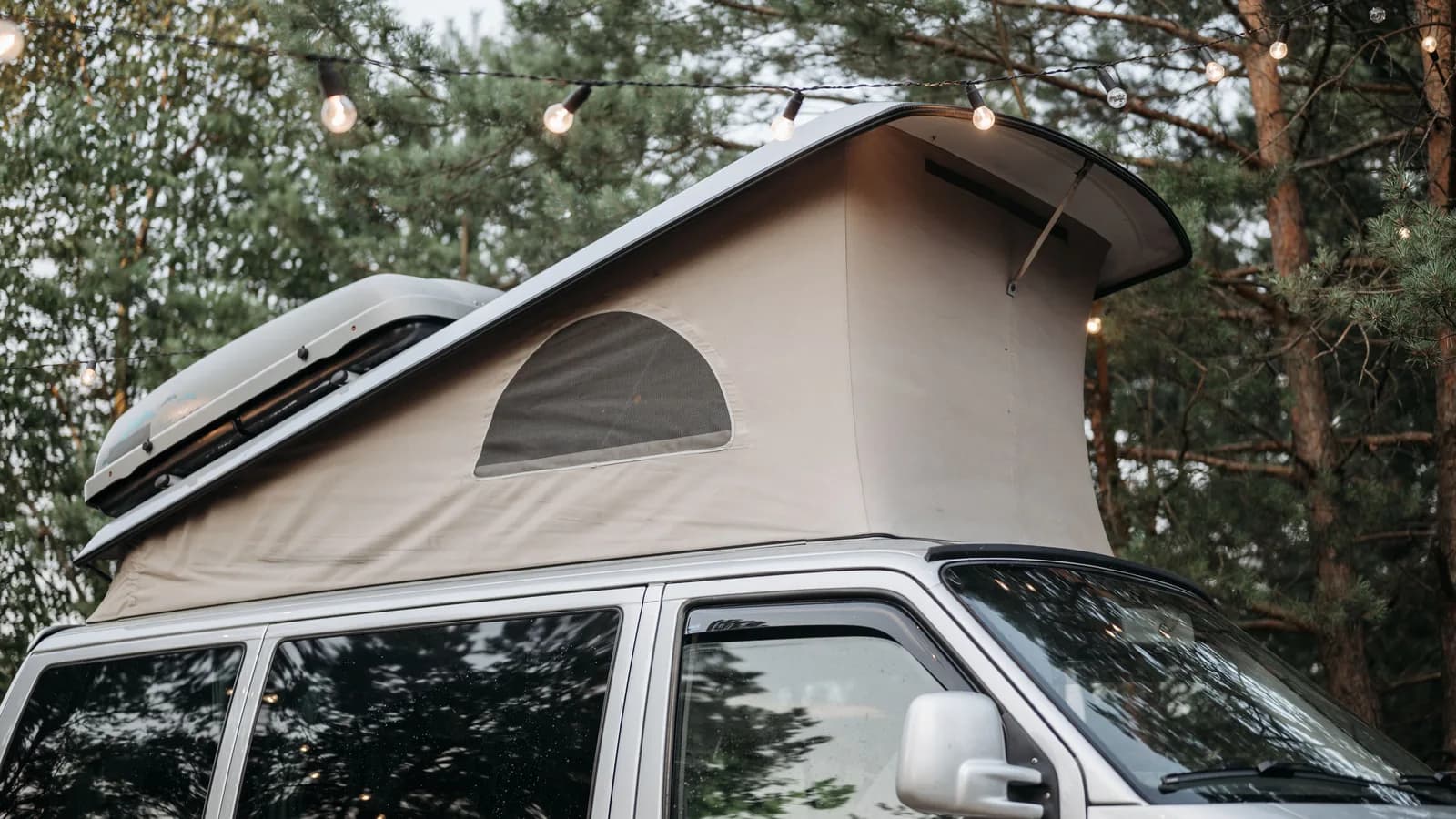 A campervan with its pop-top roof raised, parked among pine trees at dusk