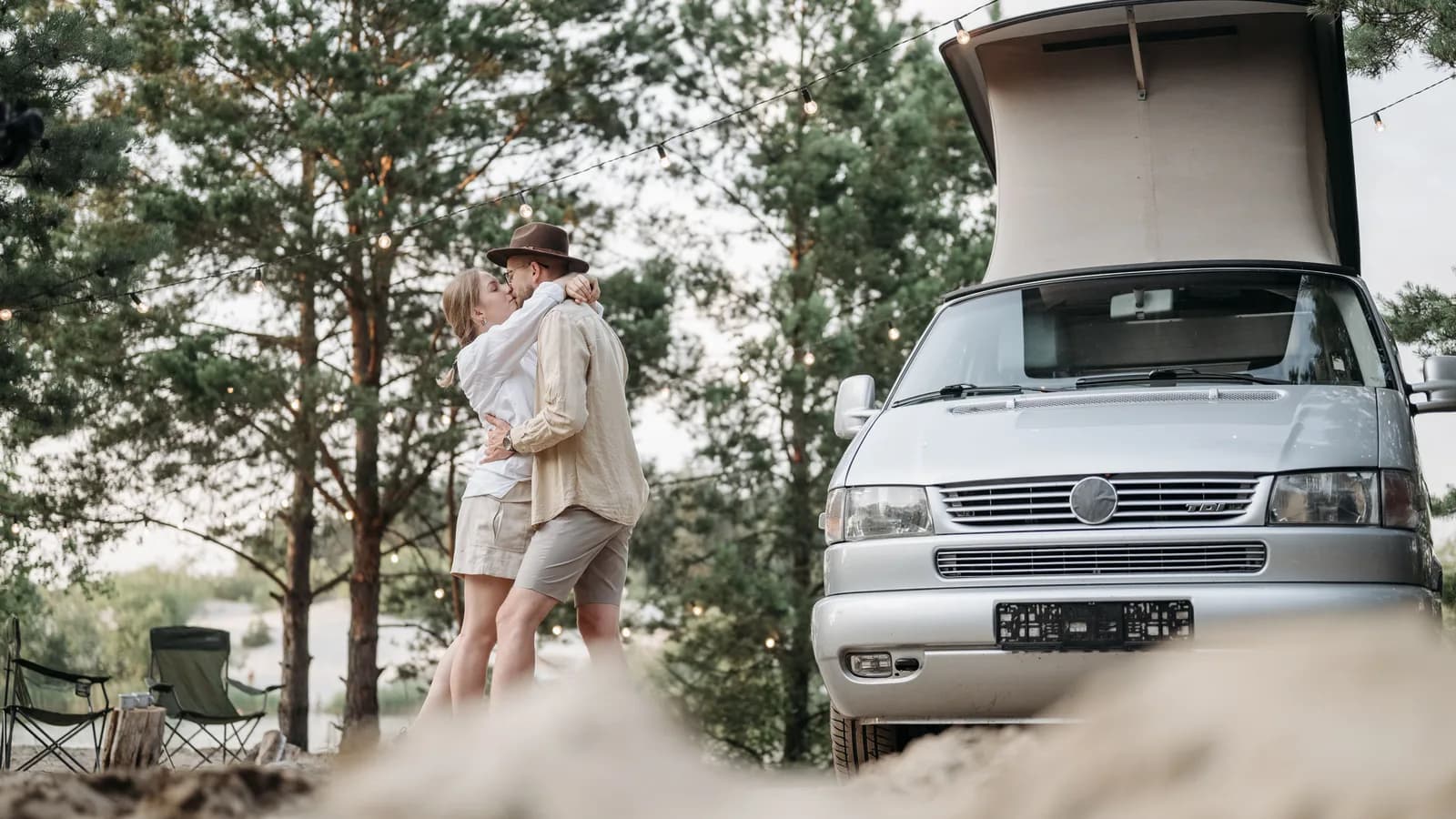 A camper van with its pop-top raised at a forest campsite, a couple standing beside it