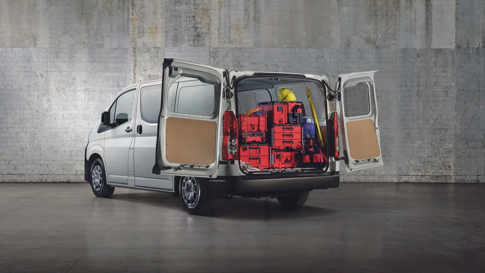 A white HiAce van with rear doors open showing red tool cases and a hard hat, against a concrete wall
