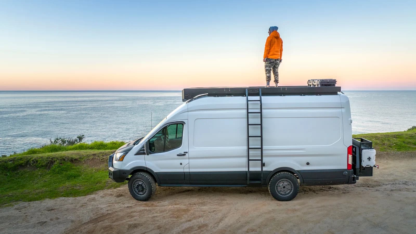 A white campervan with a roof rack and rear ladder parked on a coastal clifftop