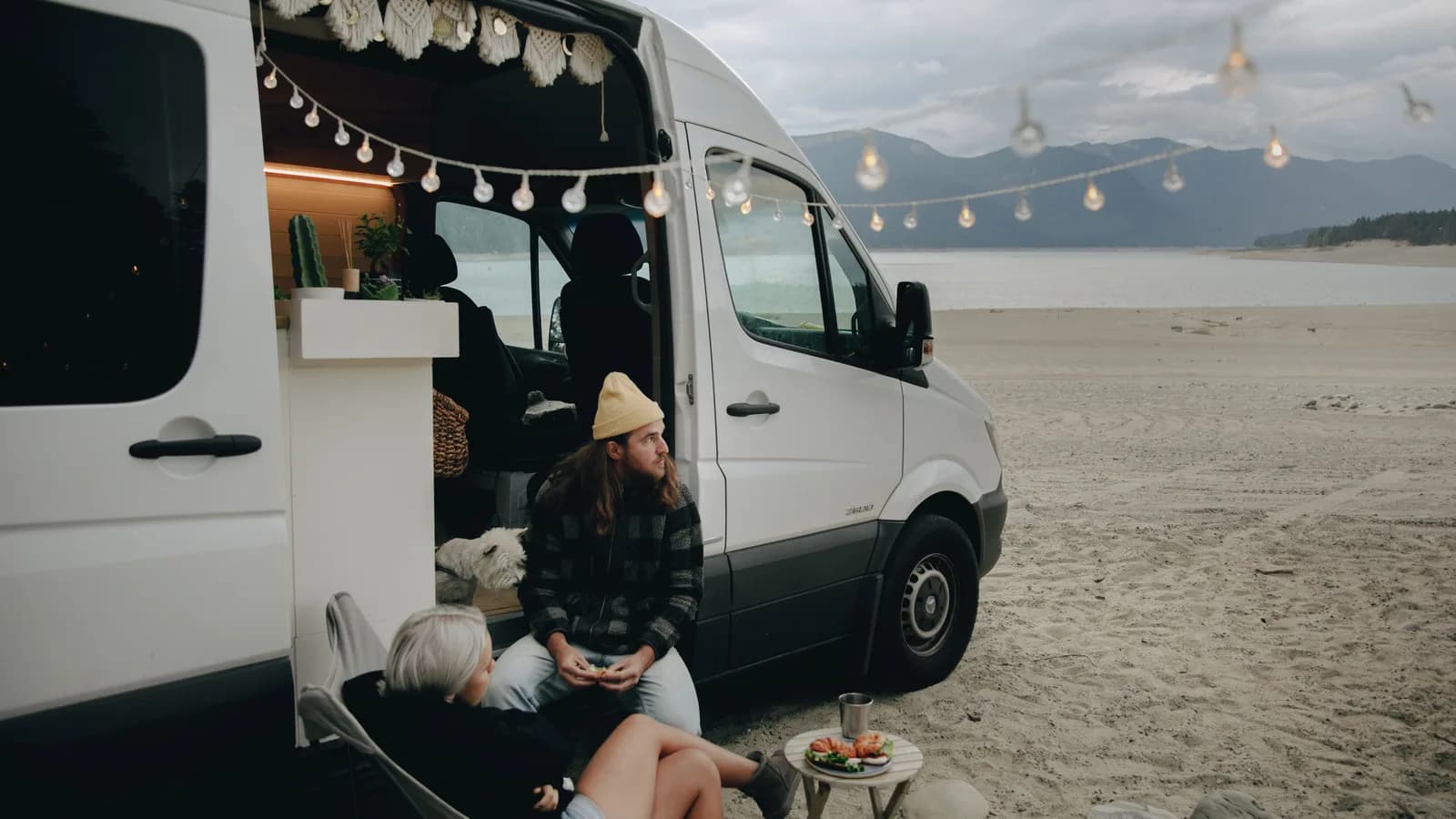 A couple sitting at the open rear doors of a campervan on a beach at dusk