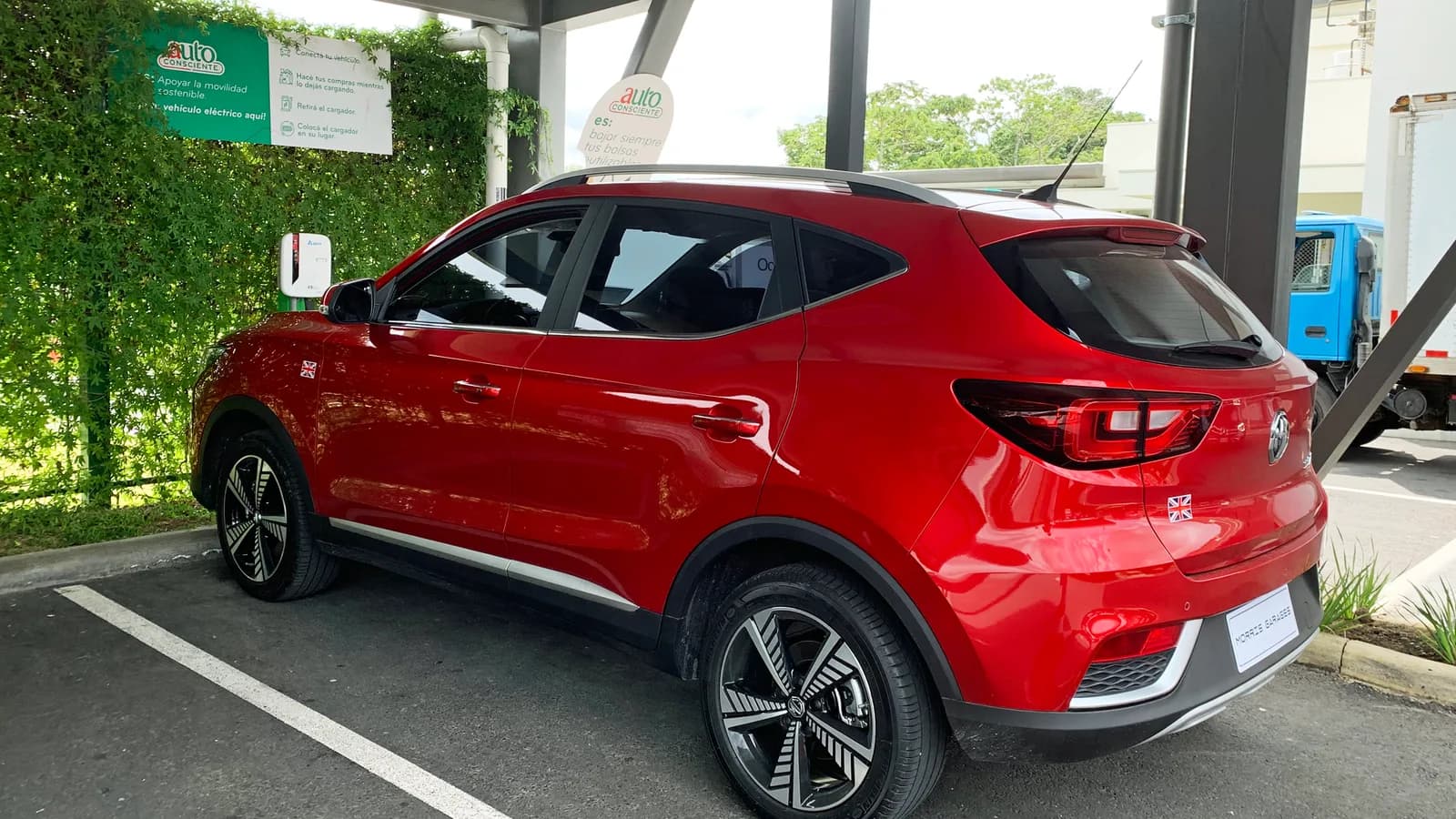 A red MG electric SUV parked beside a public charging station, viewed from the rear three-quarter