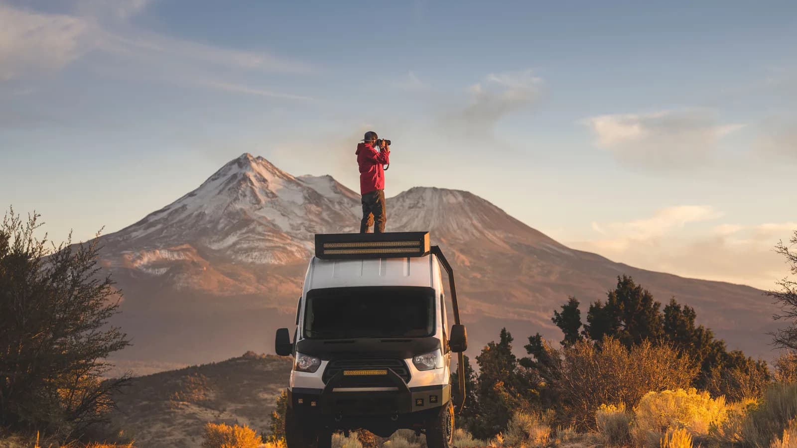 A white Ford Transit camper conversion with roof light bar and bull bar, front view, a man on the roof photographing a mountain at sunset