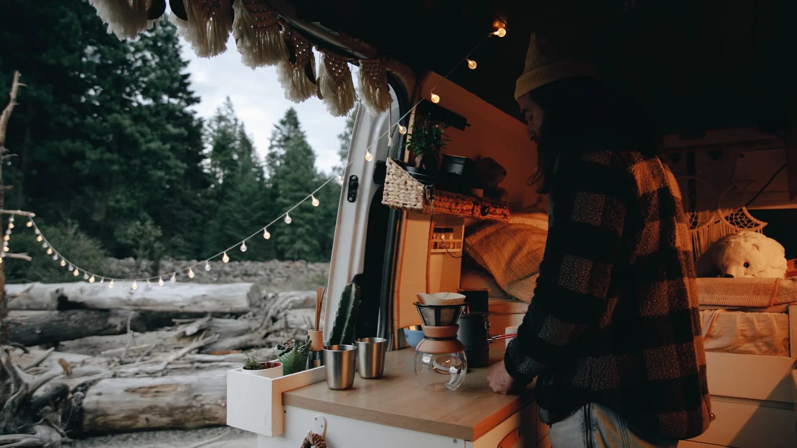 Person making pour-over coffee at a bench in the open rear of a converted van, fairy lights and bedding inside, forest and driftwood outside