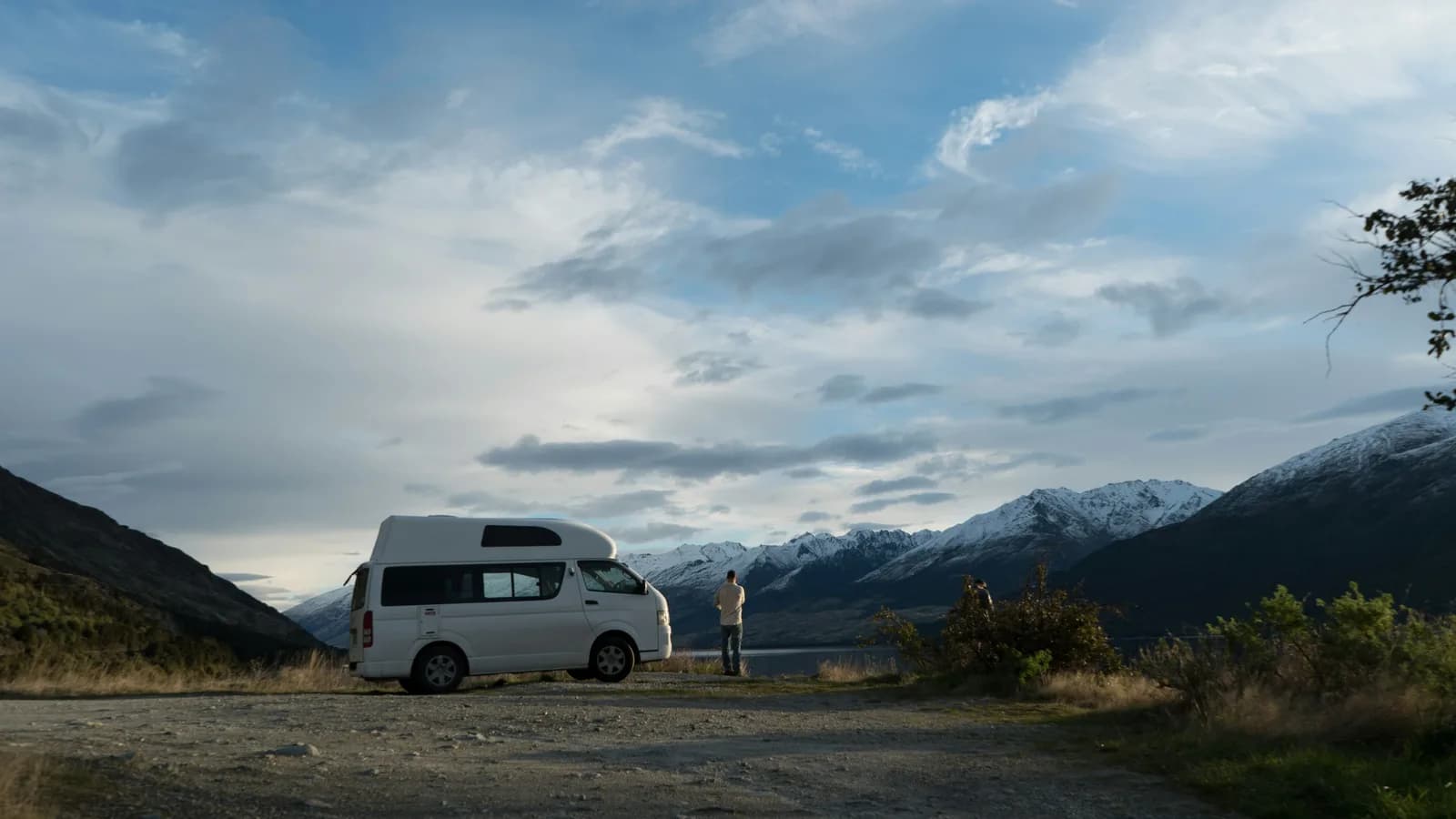 A white HiAce campervan with a raised roof parked at a gravel lookout above a lake
