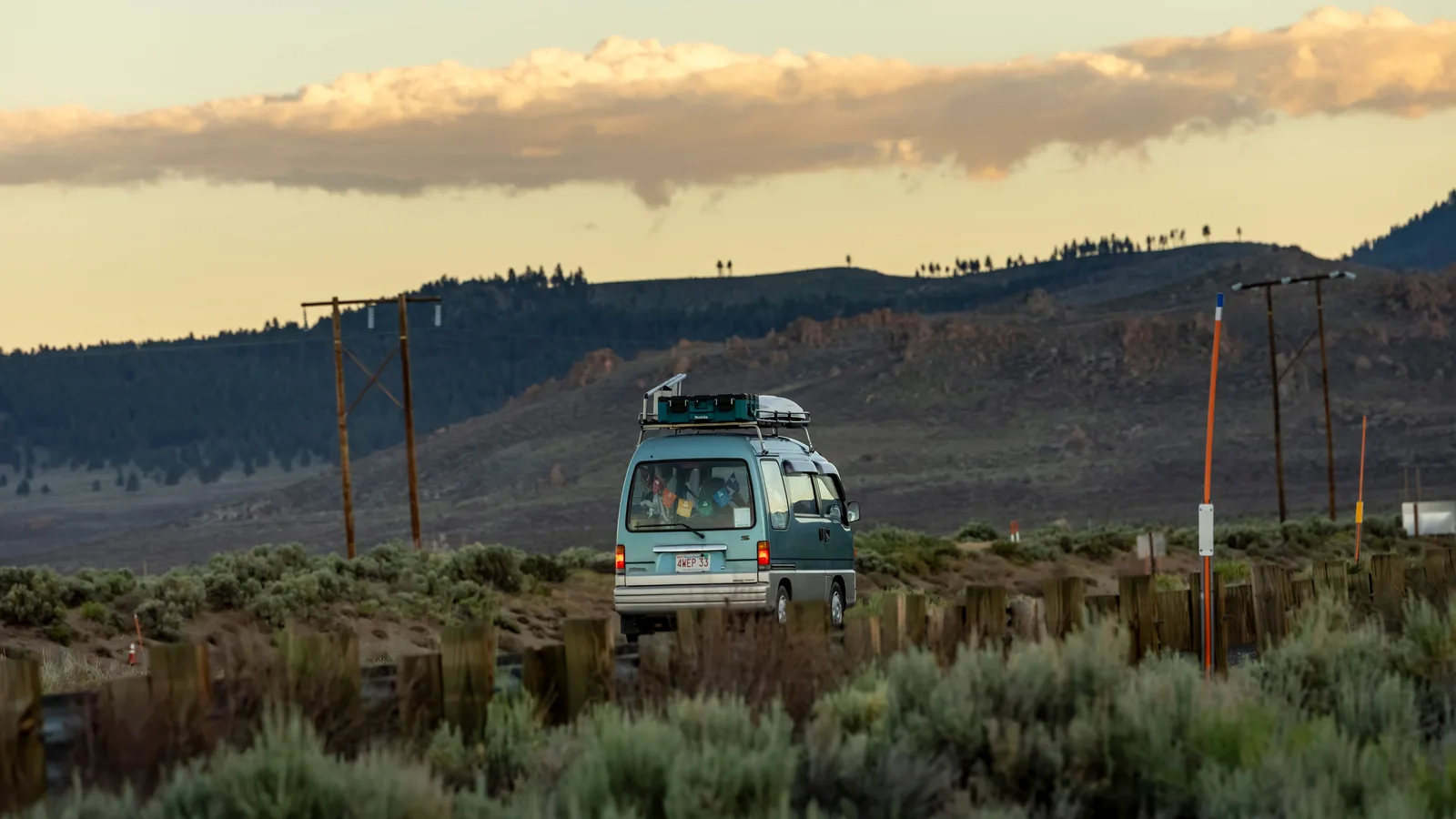 A camper van loaded with roof rack gear on a remote dirt road at dusk with mountains behind