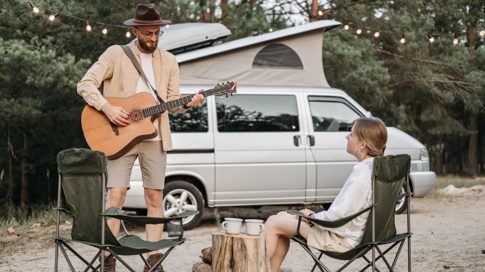 A silver pop-top camper van parked among pine trees with two people seated in camp chairs beside it