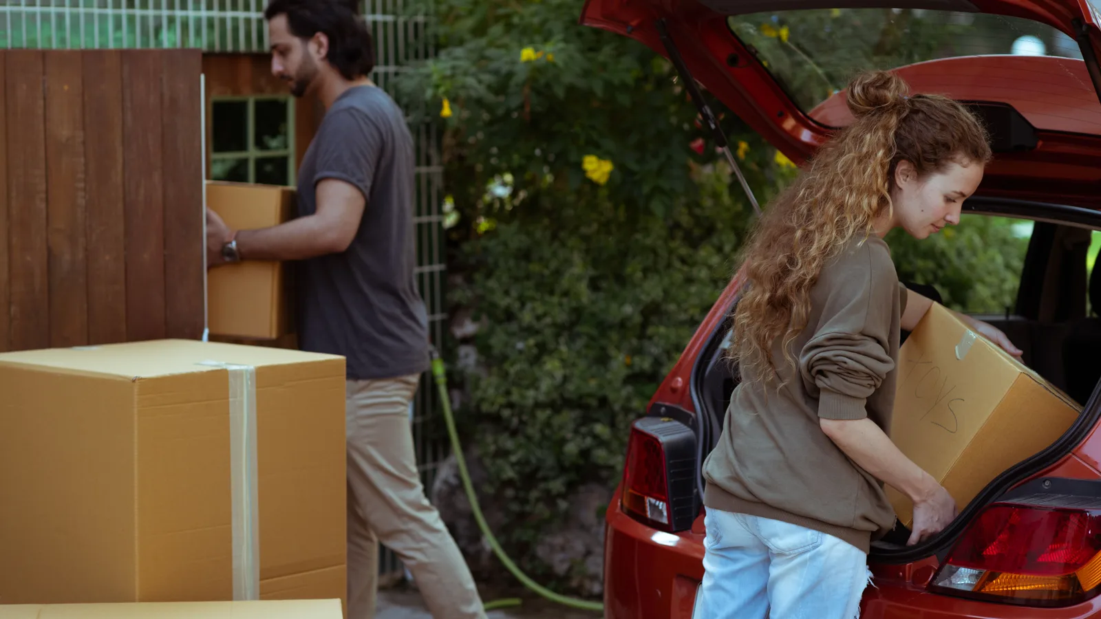 A couple lifting cardboard boxes into the open boot of a red hatchback in a driveway