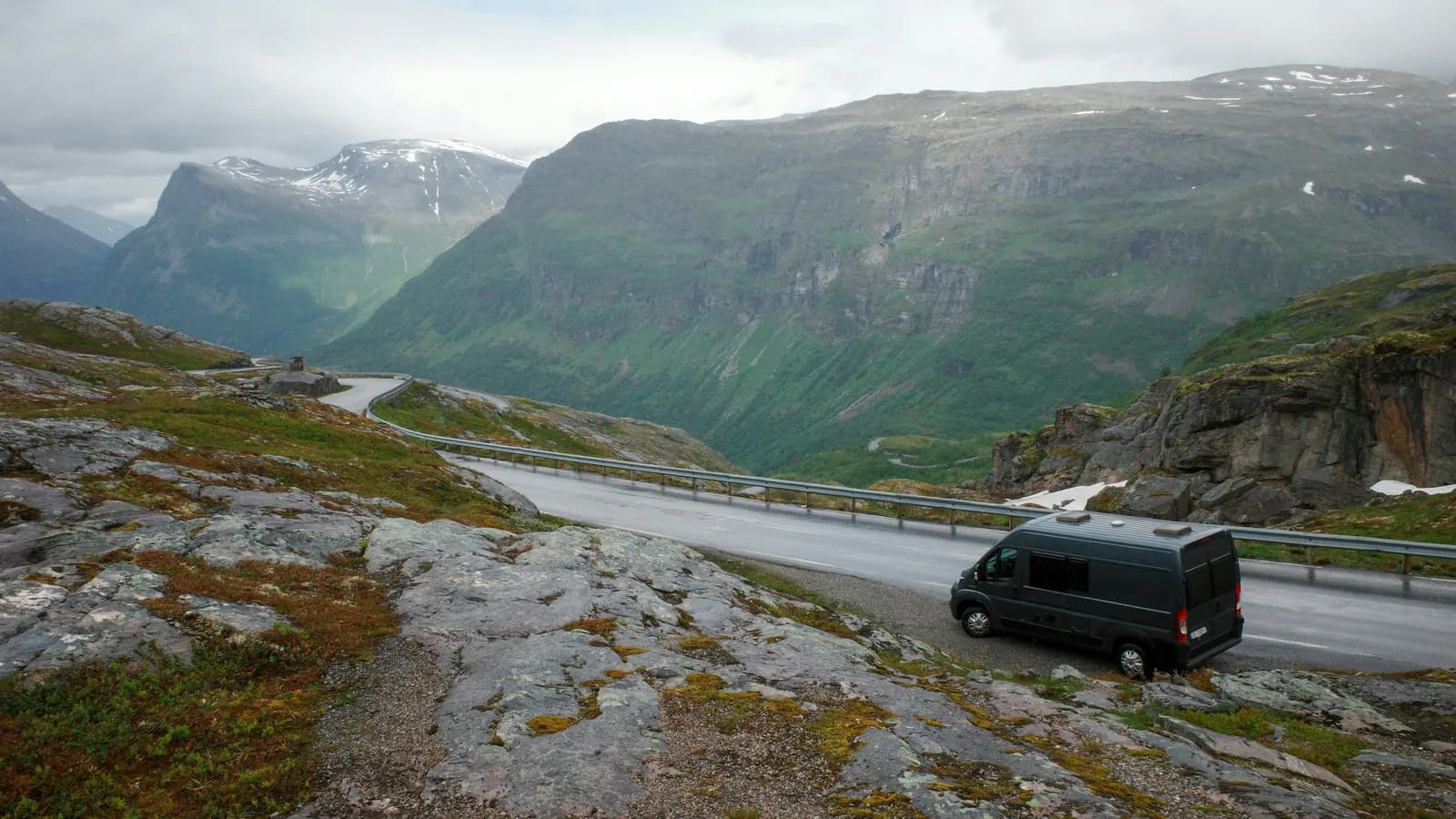 A dark grey panel-van camper parked at a lay-by beside a guard-railed mountain road with a valley behind, aerial view