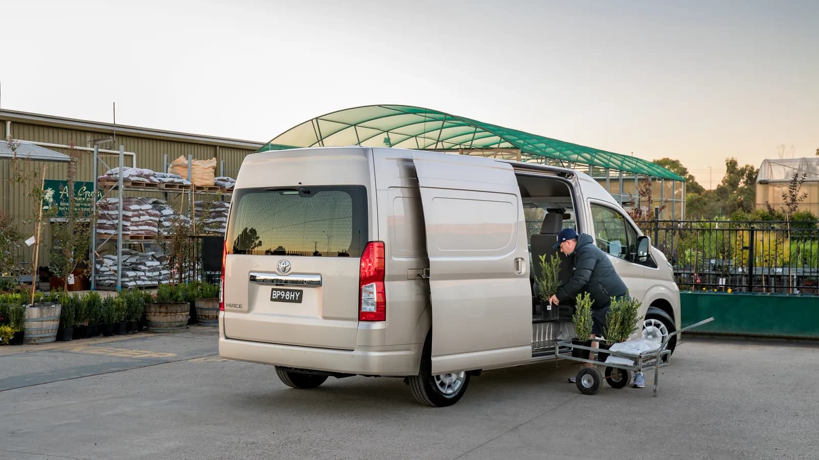 A beige Toyota HiAce van with Australian plates, rear three-quarter view, sliding door open at a plant nursery while a man loads potted plants