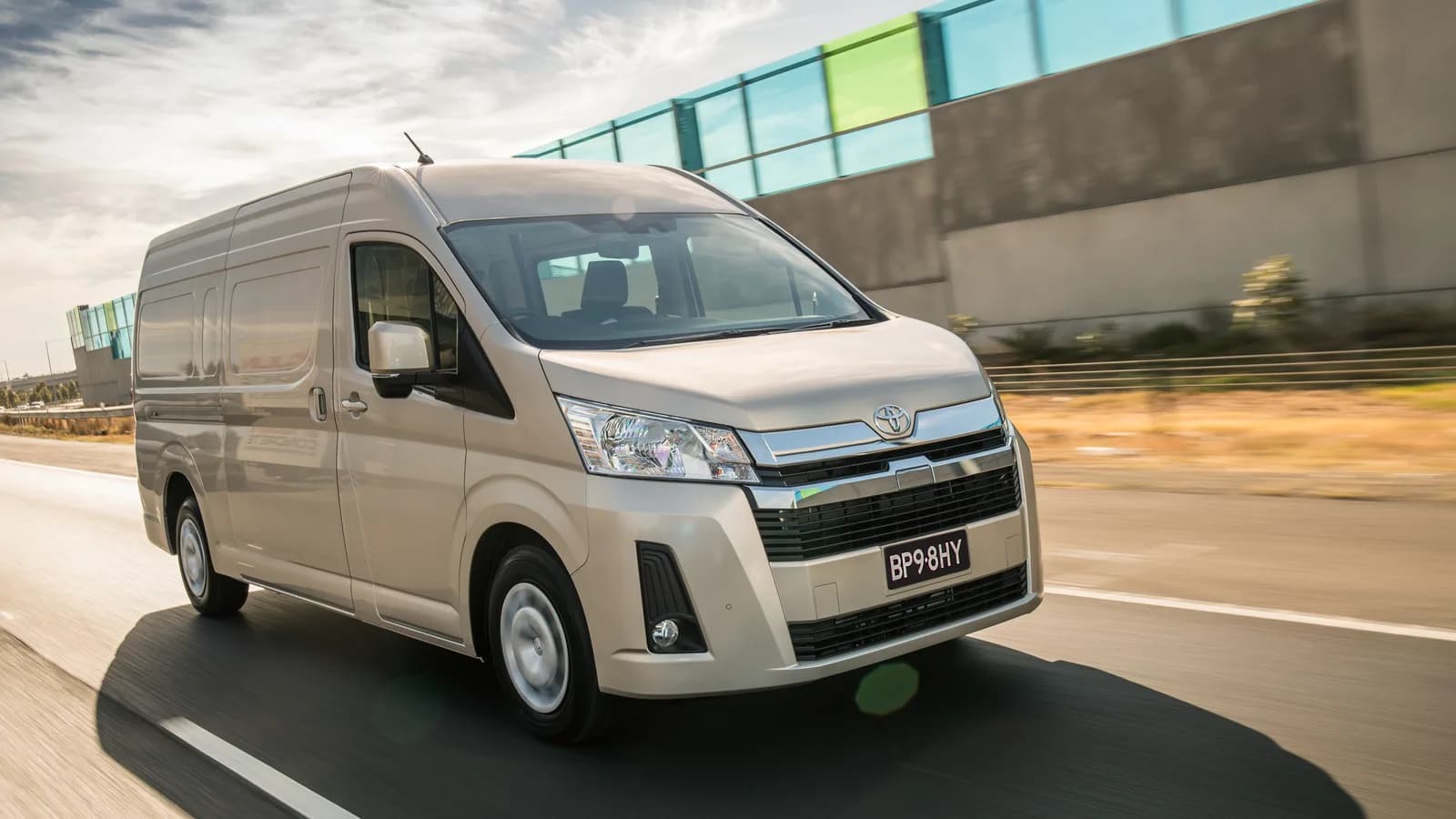 A beige high-roof Toyota HiAce van driving on a motorway, three-quarter front view