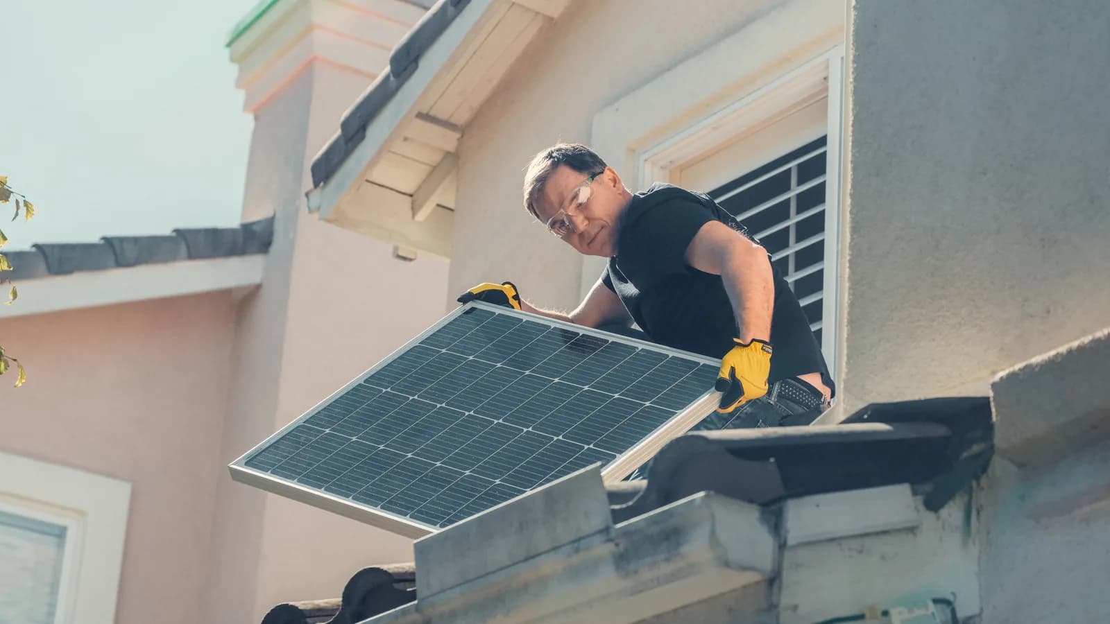 A man in work gloves lifting a solar panel into position along a roof edge