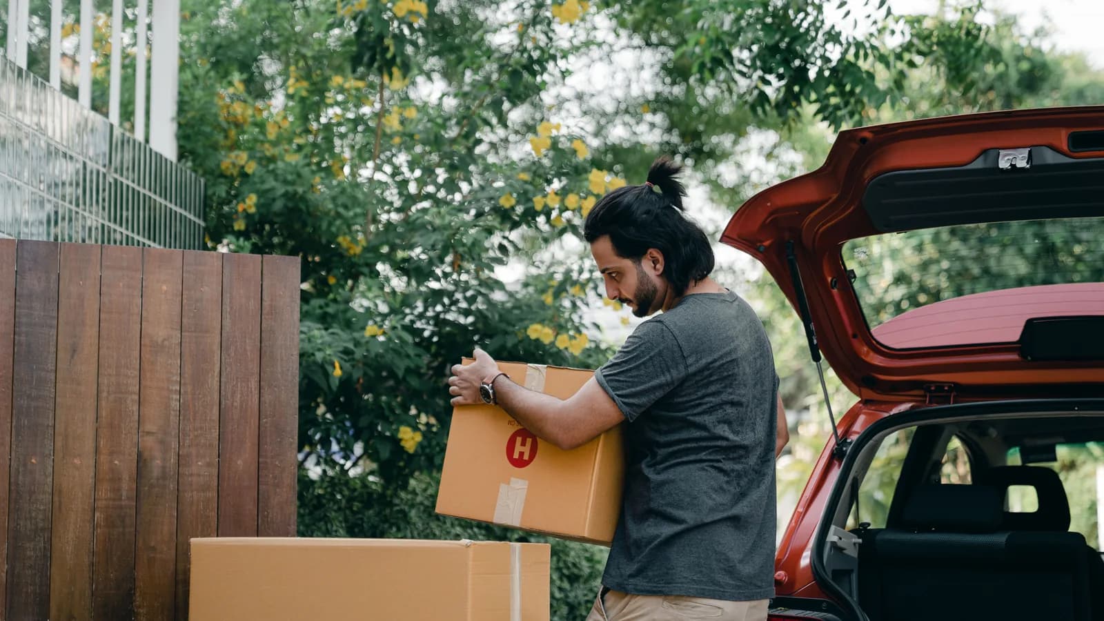 A man carrying a large cardboard box towards the open boot of a red car