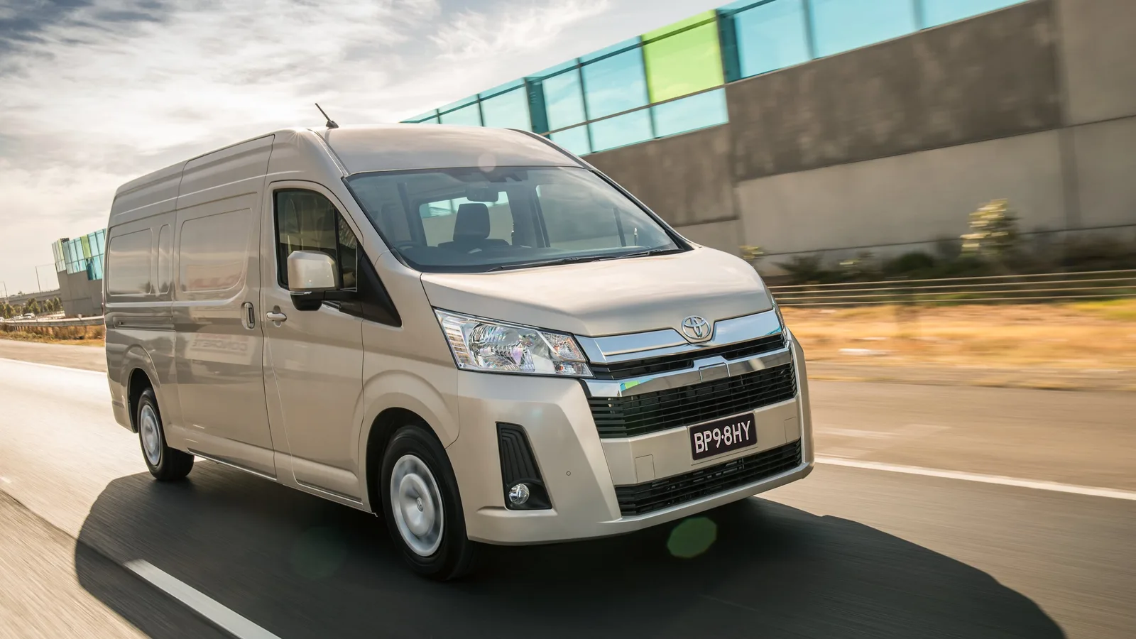 A beige high-roof HiAce van driving past a building, front three-quarter view