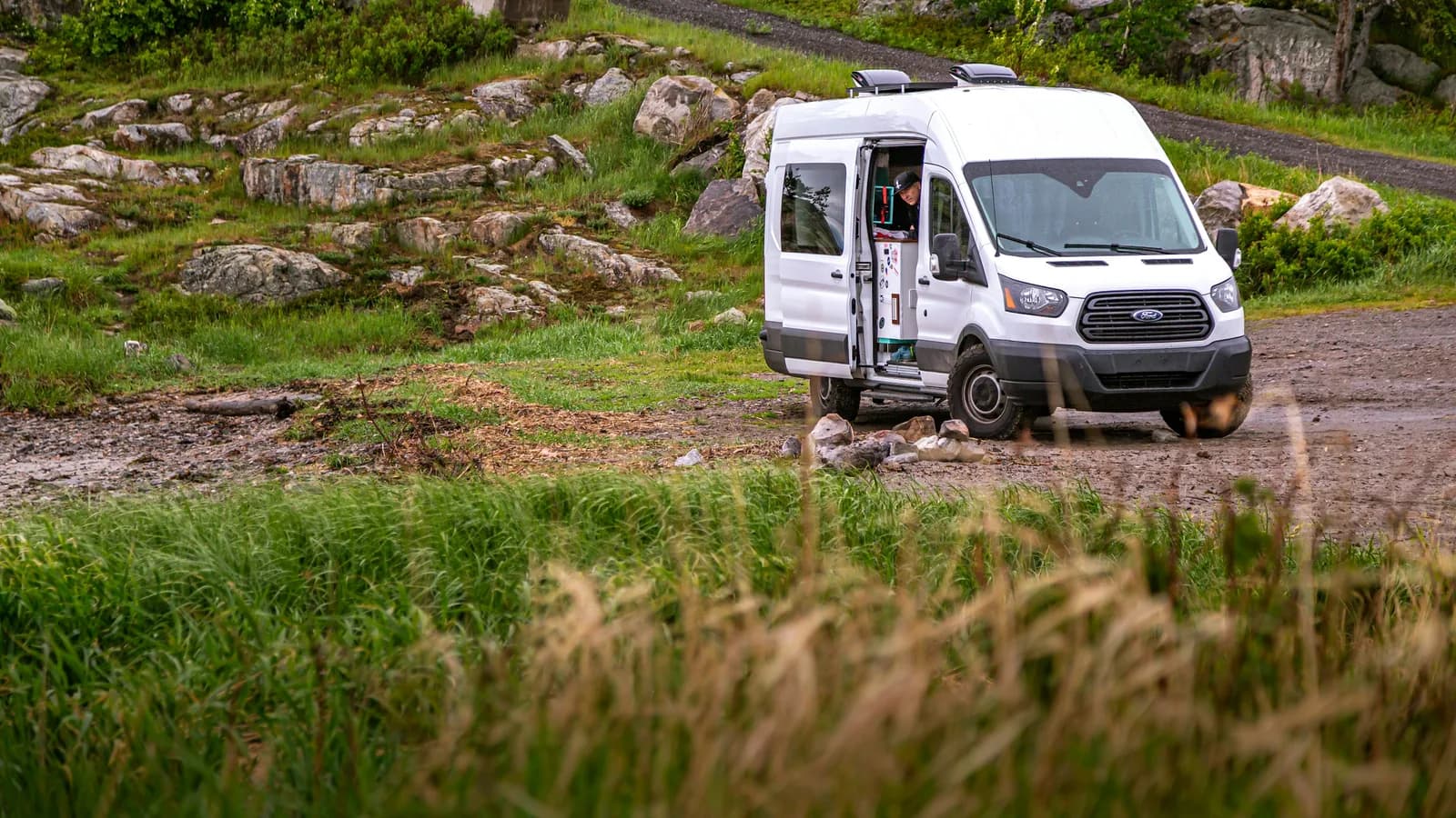 A white Ford Transit campervan with its side door open, parked on a rocky bush track