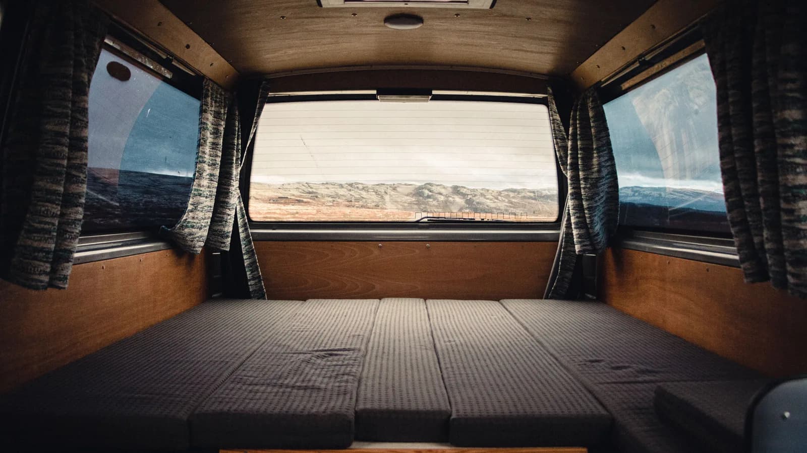 The inside of a converted van with a flat bed platform, timber wall lining and curtains at the rear windows