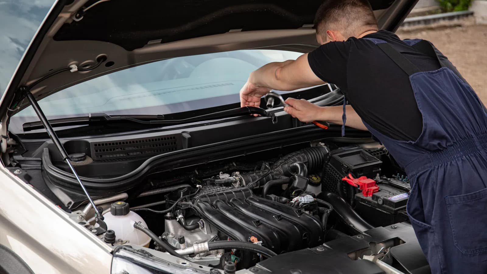 A mechanic in overalls working with a wrench in the open engine bay of a white car, outdoors