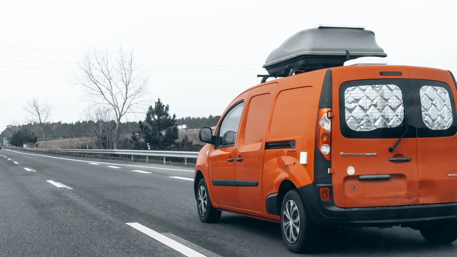 An orange van on a highway with reflective foil covers fitted inside its rear windows and a box on the roof
