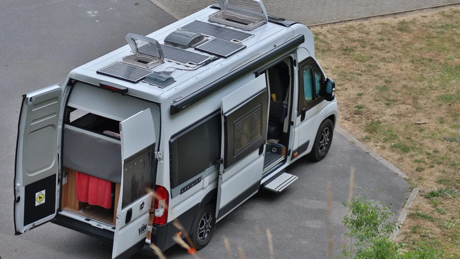 A white camper van with its rear doors open showing a plywood interior, with solar panels visible on the roof