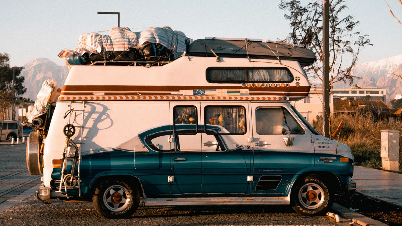 A teal and white camper van parked at a kerb with luggage strapped to its roof rack and mountains behind