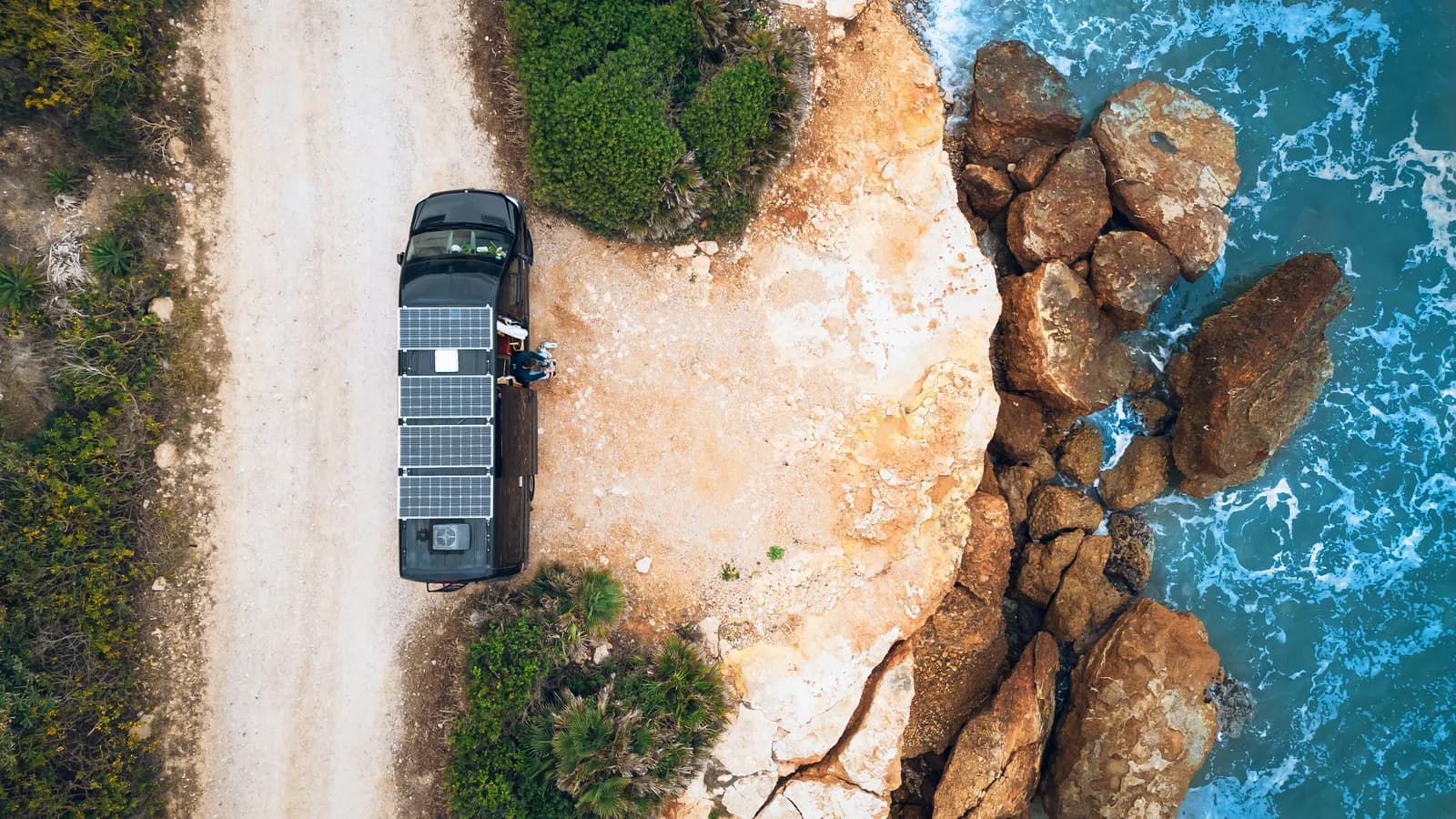 Aerial view of a camper van with roof-mounted solar panels parked on a dirt track beside a rocky coastline