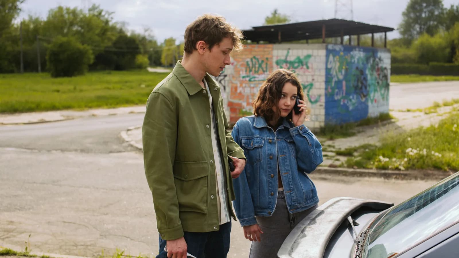 Two people standing beside a car, one on the phone, talking about a sale