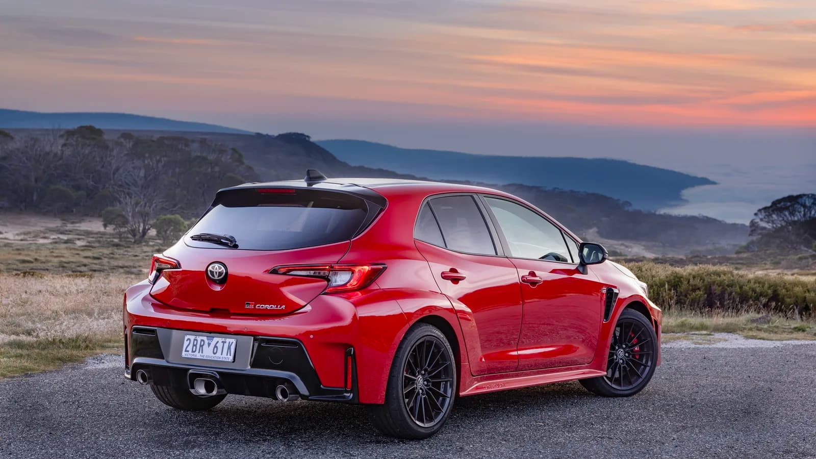 A red hatchback parked on a coastal overlook at sunset, seen from the rear three-quarter angle