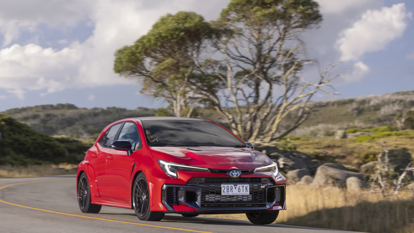 A red hatchback driving along a rural road with hills and trees behind it