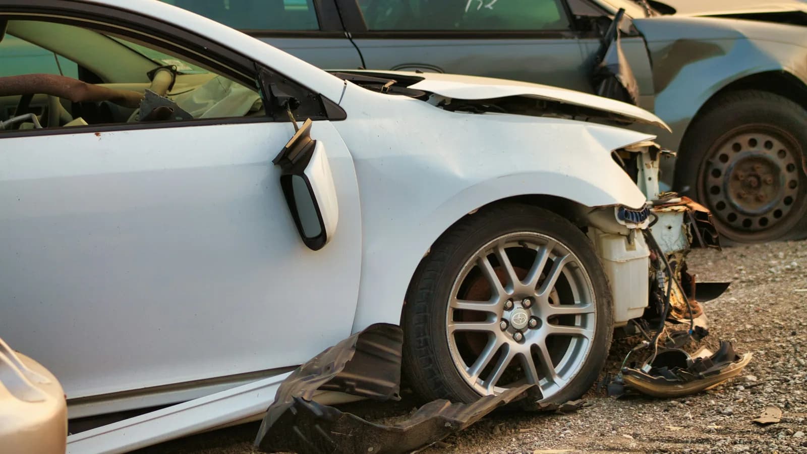 A white car with a smashed front end and crushed wheel arch sitting in a salvage yard