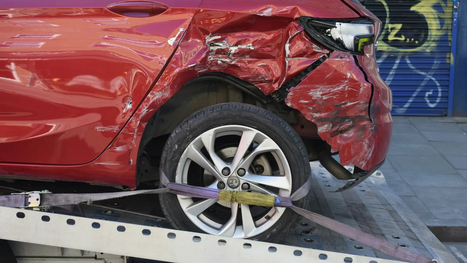 A damaged red hatchback strapped to the tray of a flatbed tow truck