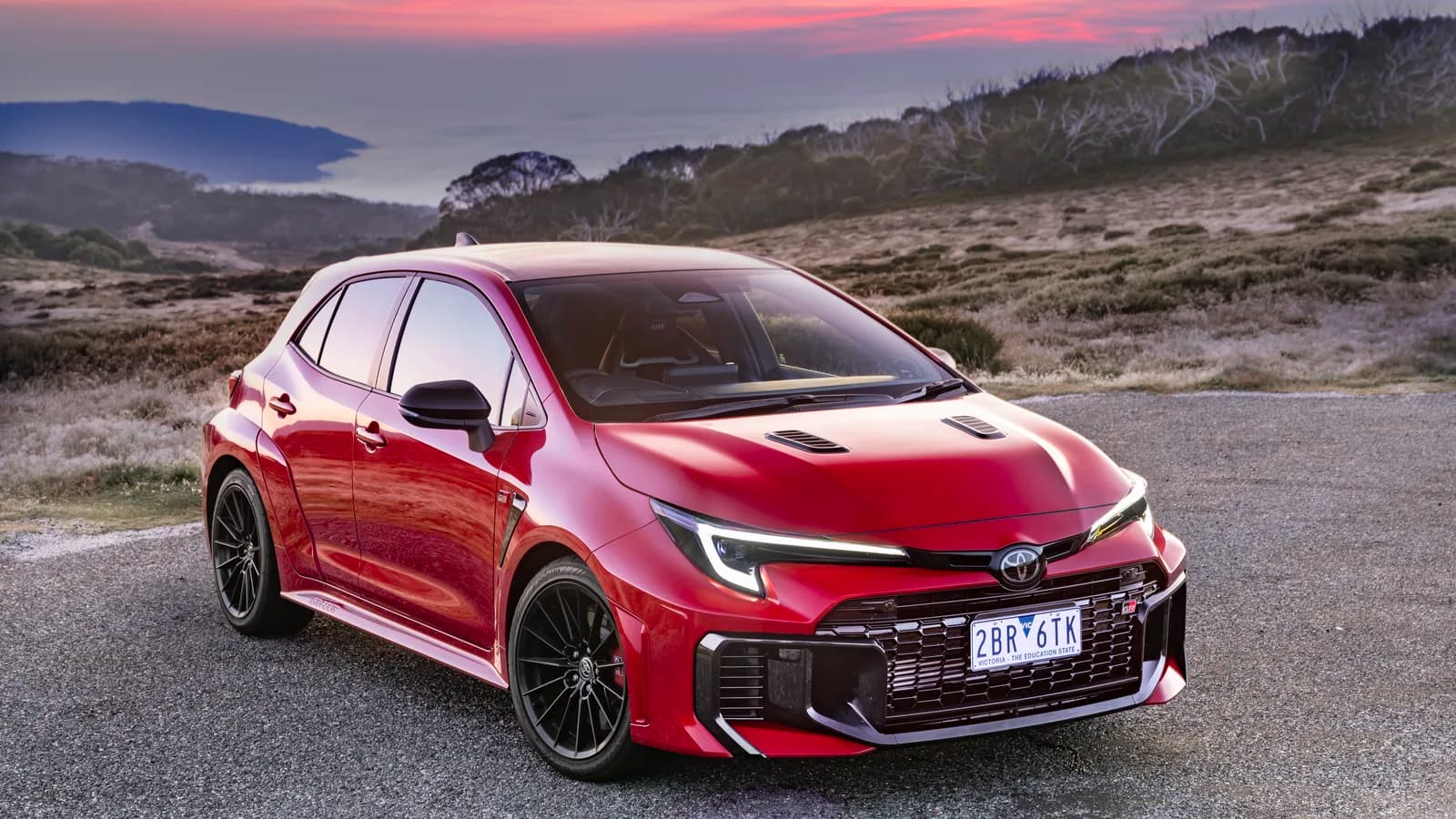 A red hatchback parked on a gravel overlook at sunset, seen from the front three-quarter angle