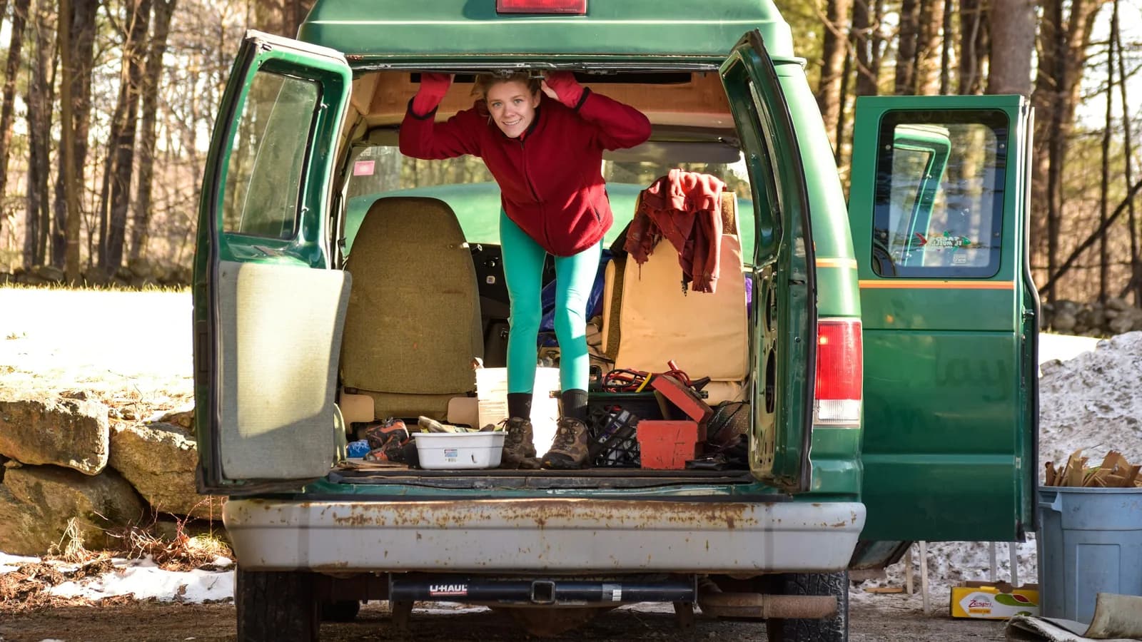 A woman standing in the open rear load area of a converted van packed with bedding and gear