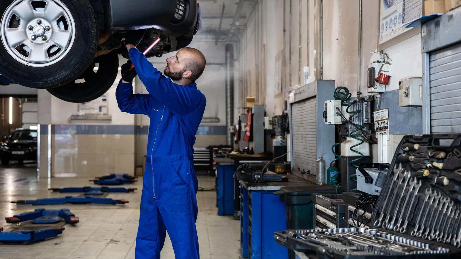 A mechanic in blue overalls looking up at the underside of a van raised on a workshop hoist