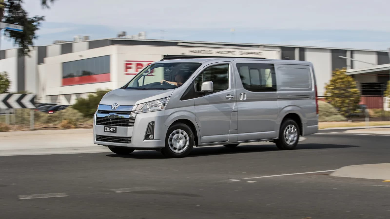 A silver Toyota HiAce van passing a large industrial building