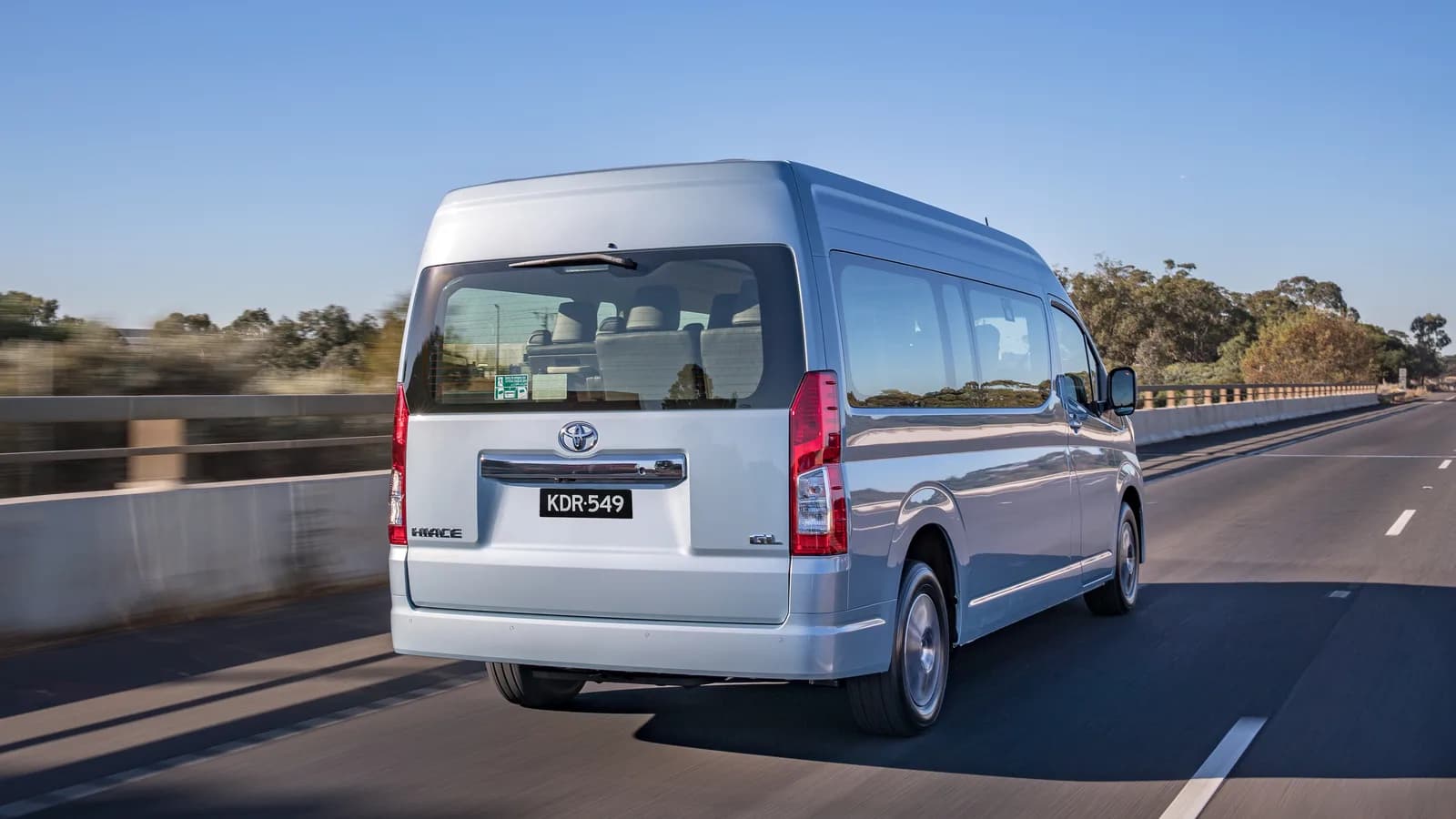 A silver Toyota HiAce driving away along an open road under a clear sky