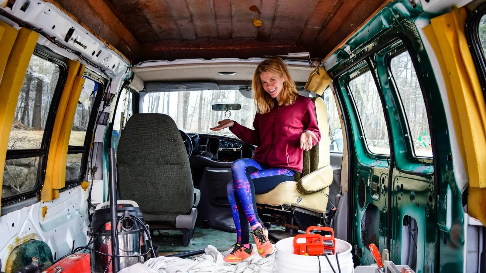 A woman sitting inside a stripped-out van part way through a camper conversion, tools on the bare floor