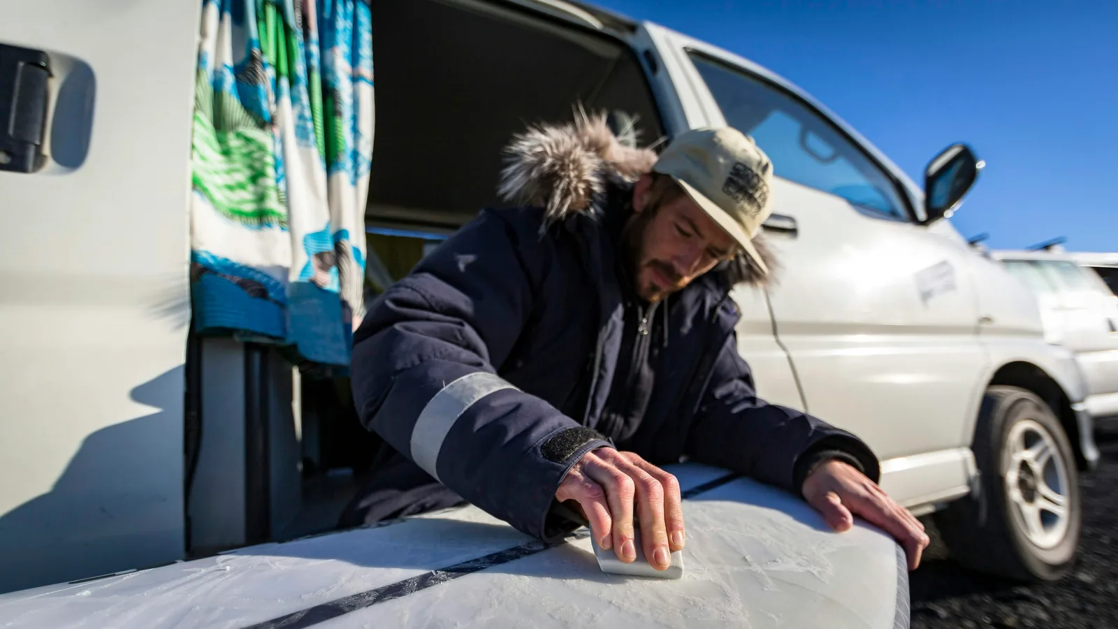 A man waxing a surfboard beside a van fitted out for camping, curtains drawn in the side window