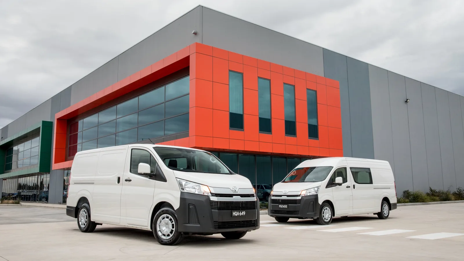 A short-wheelbase and a long-wheelbase white HiAce parked side by side outside a commercial building