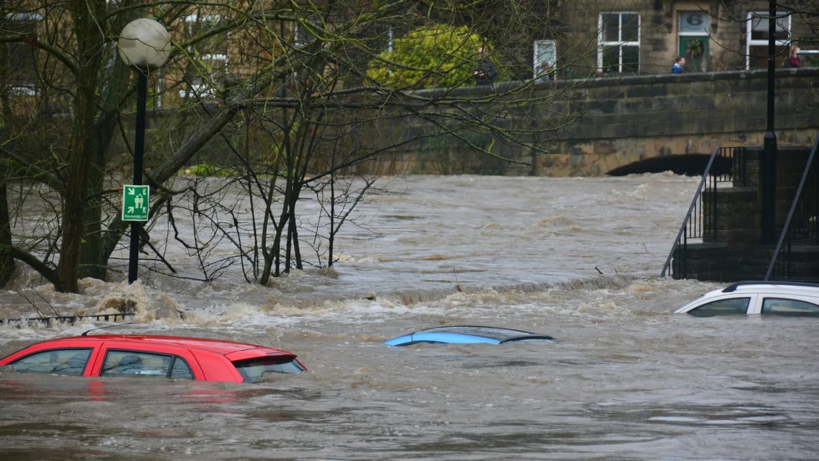 Two cars almost fully submerged in brown floodwater next to a bridge