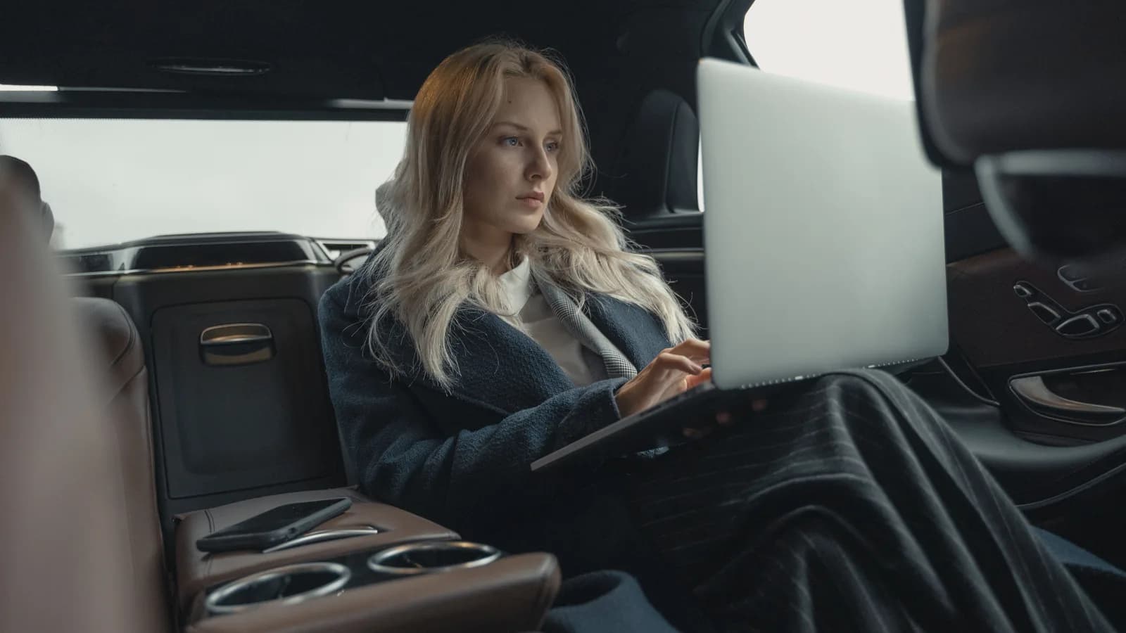 A woman in a blazer sitting in the back seat of a car typing on a laptop